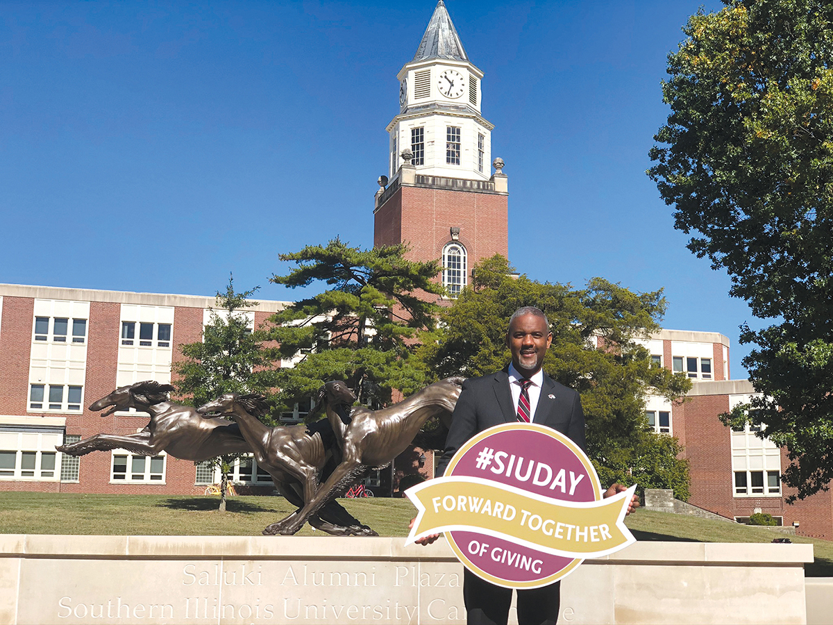 Chancellor Austin A. Lane holds SIU Day of Giving 2021 sign in front of the Saluki Alumni Plaza and Pulliam clocktower.